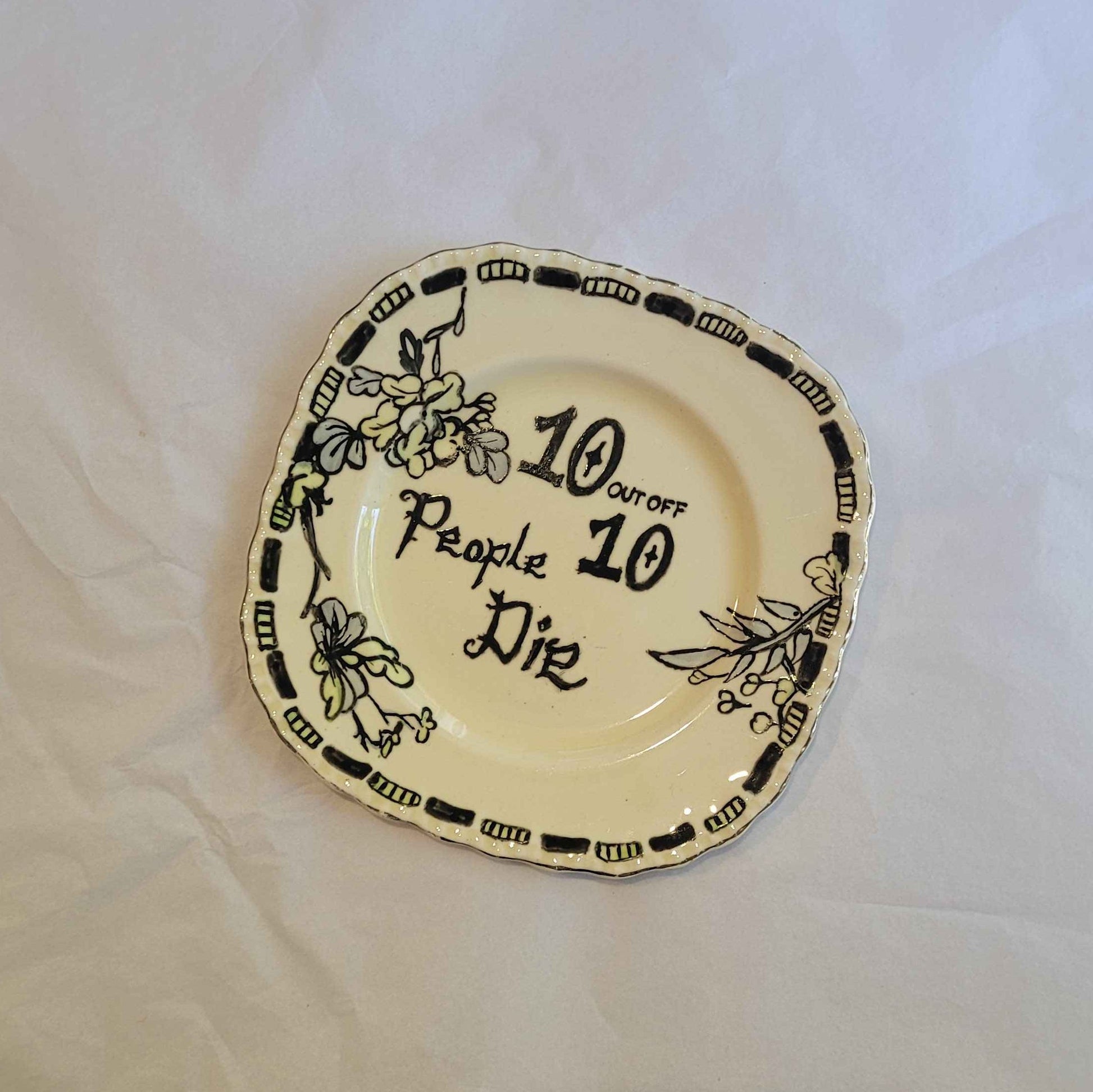 a small porcelain plate with hand pained ink detailing. the plate features the words "10 out of 10 people die" however of is misspelled as "off". the design also features a dashed boarder around the edge and some floral elements. 