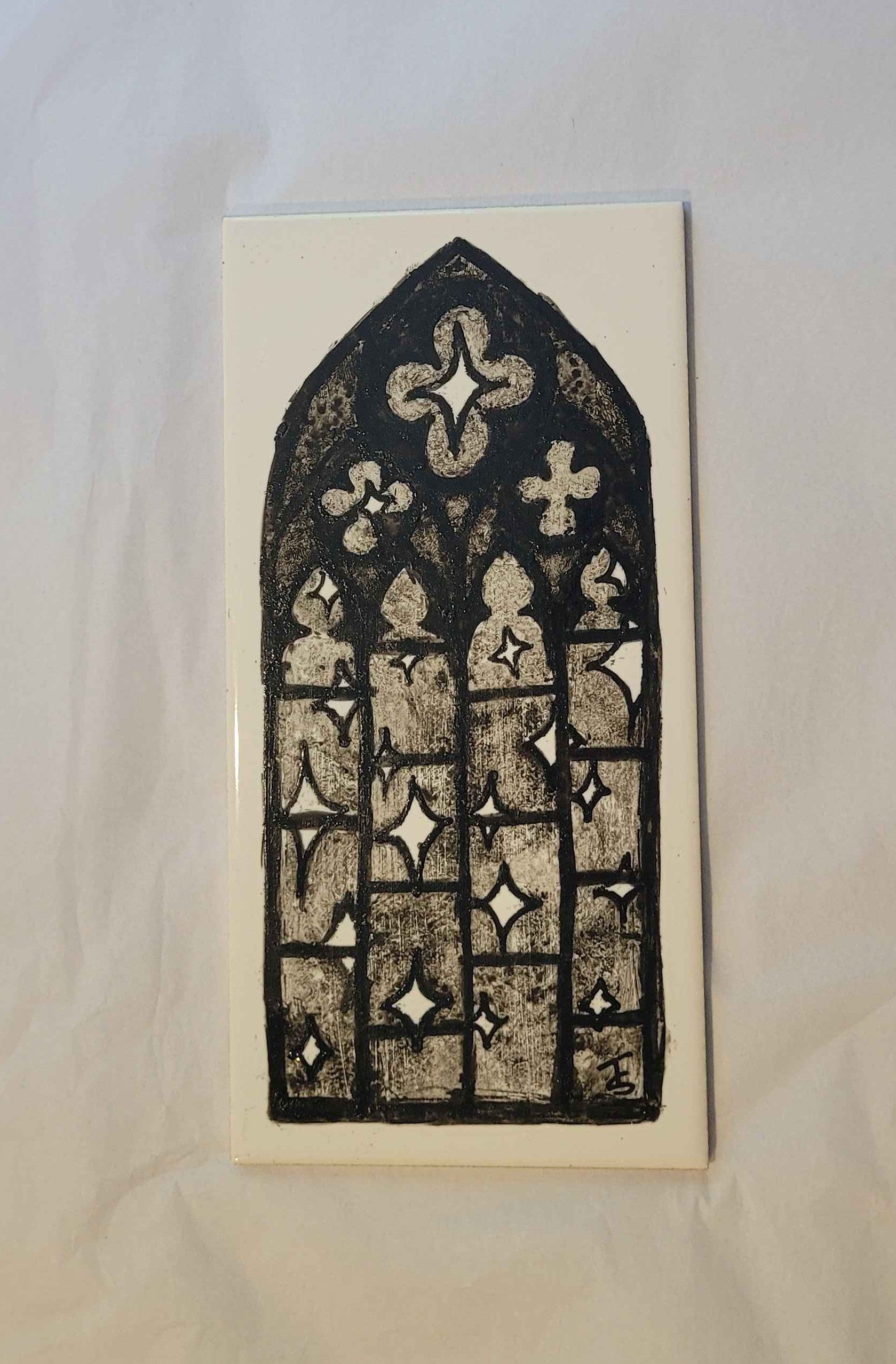 A ceramic tile, featuring a hand-painted ink illustration of a gothic style window. in the window you can see a dark night sky full of 4 pointed whit stars. 