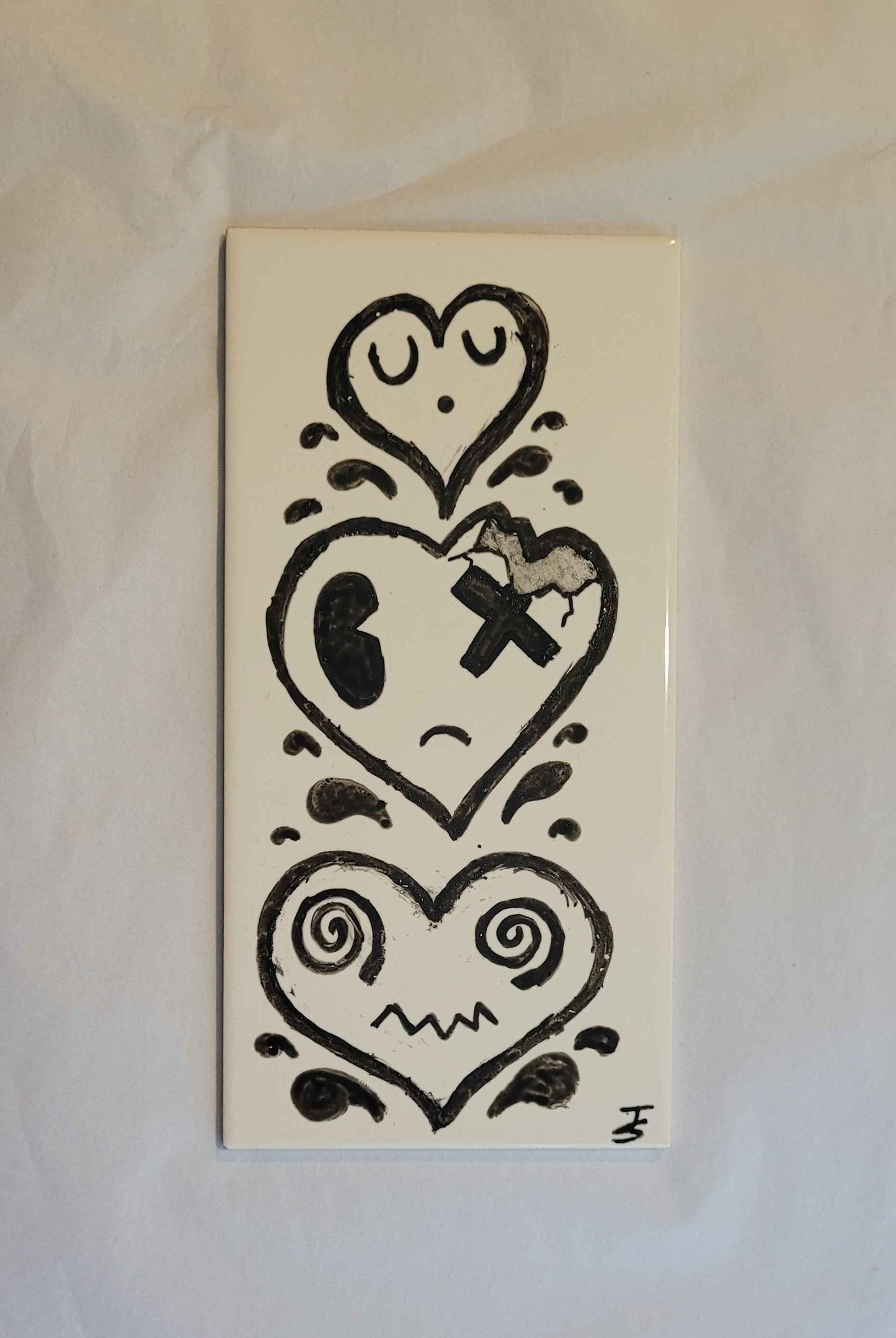 A ceramic tile featuring a hand painted ink illustration of 3 cartoon hearts with faces and droplets between them. 
the top heart is small and sleeping. 
the middle heart is carked and is missing the top right corner, the eye on that side is an x and it is frowning. 
the bottom heart is wider, with swirls for eyes and a zigzag mouth. 