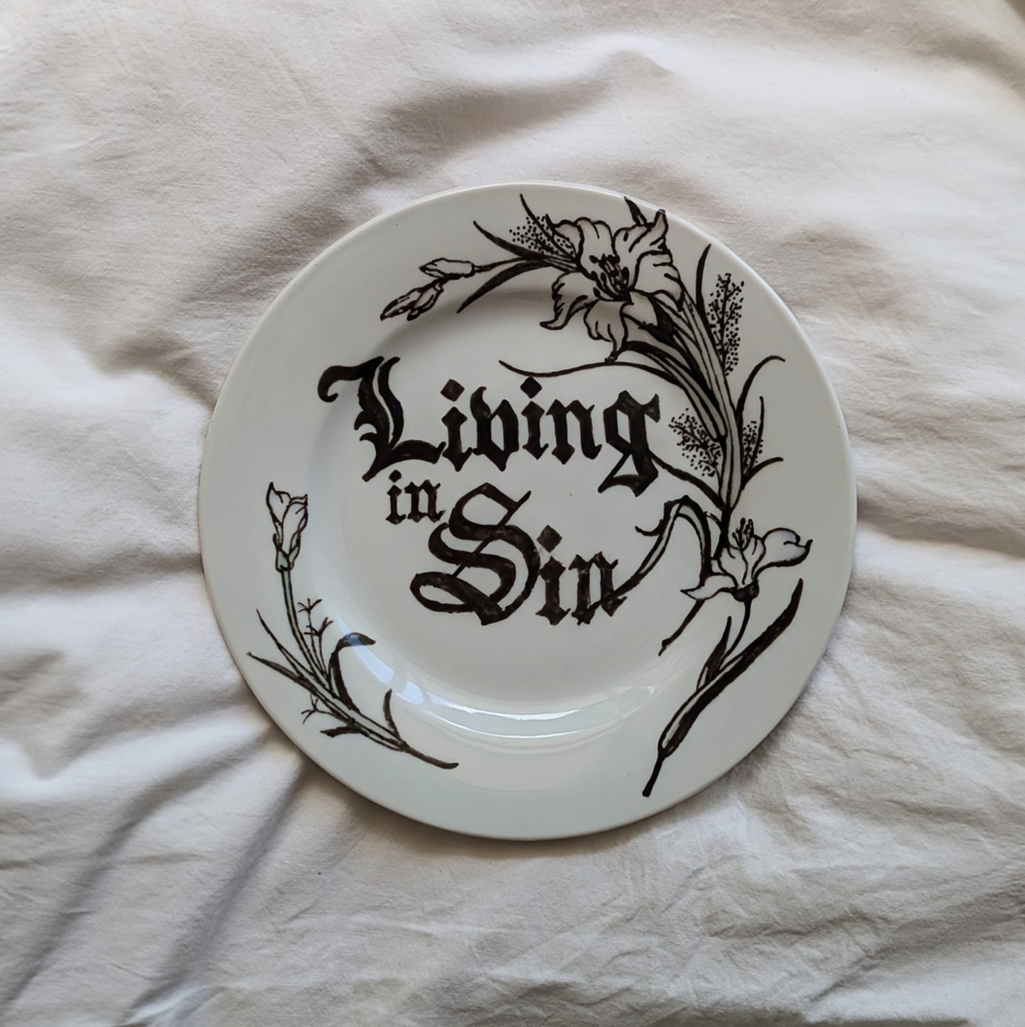 A small plate with a black out lined flora design. It features the words "living in sin" hand written in the centre in a black letter font.