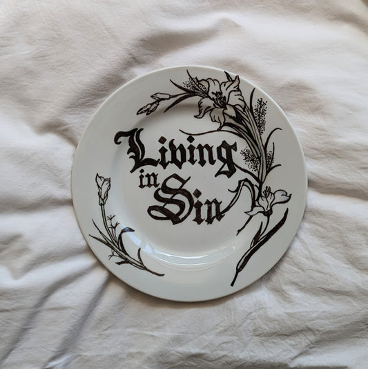 A small plate with a black out lined flora design. It features the words "living in sin" hand written in the centre in a black letter font.