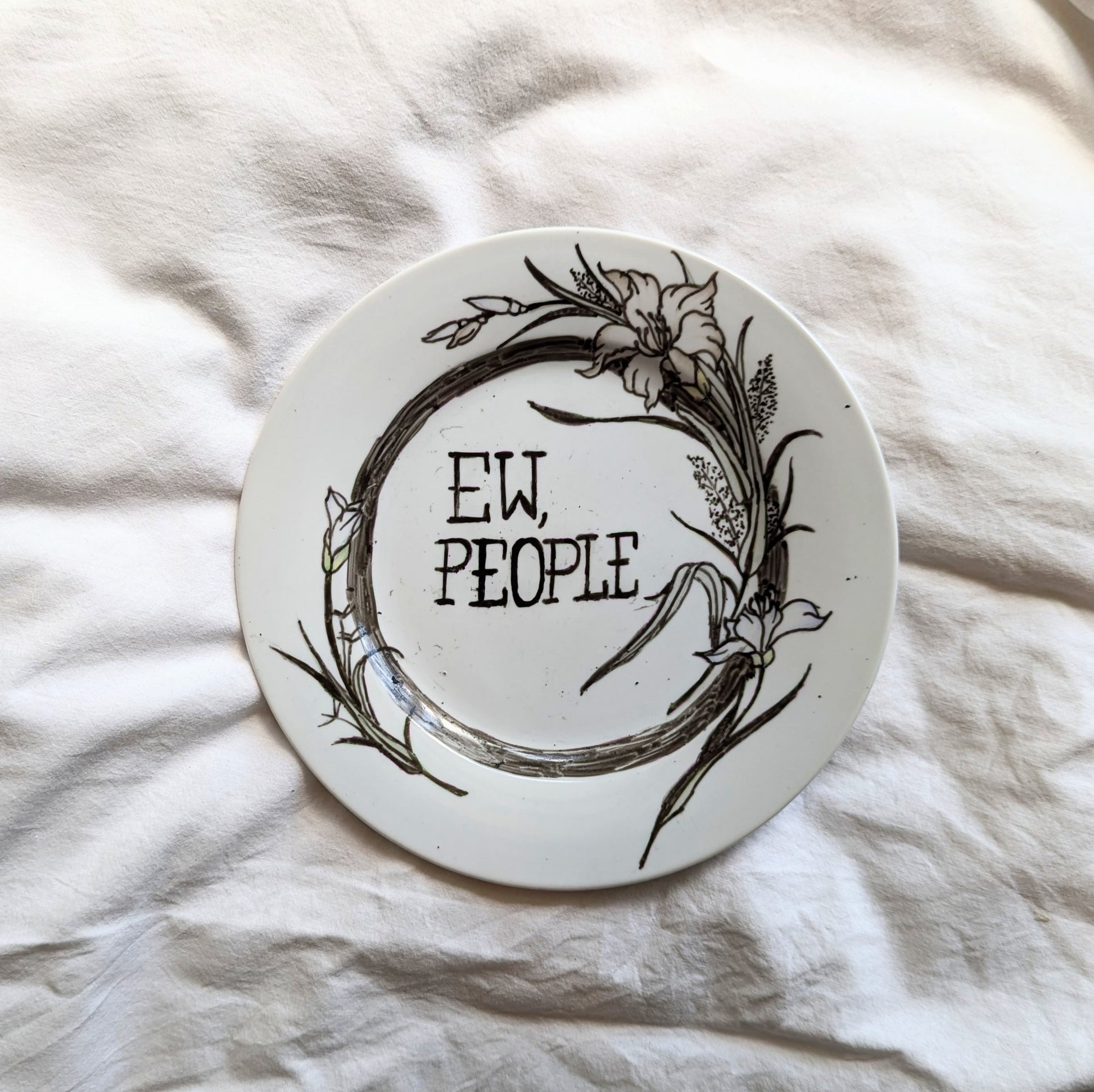 A small side plate with black outlined flora prints. In the centre are the words "ew people" hand written in a cross stitch like font.