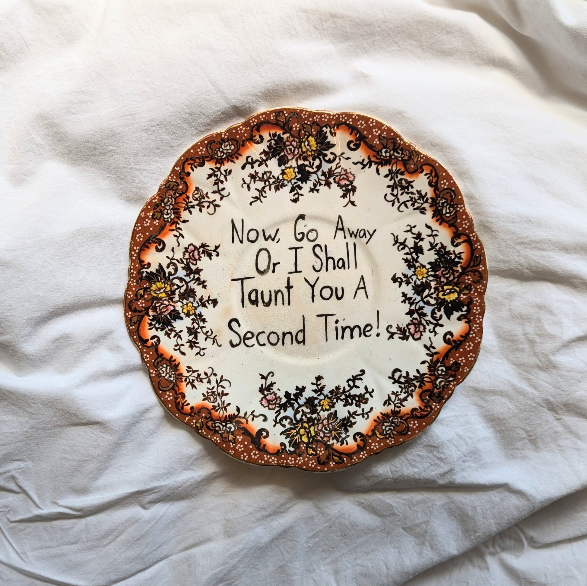 A highly detailed antique looking plate with red gold and flora designs around the rim. In the centre of the plate are words written "now go away or I shall taunt you a second time" in ink.