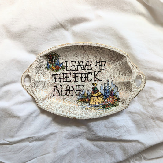 A decorative platter featuring a flora all over gold design with two sections of coloured flowers in the top left and bottom right corners. In the right corner There is a woman walking through a garden in a yellow old timey Bonet and dress. The the middle of the plate in black ink are the words "leave me the fuck alone" in a handwritten cross stitch style.