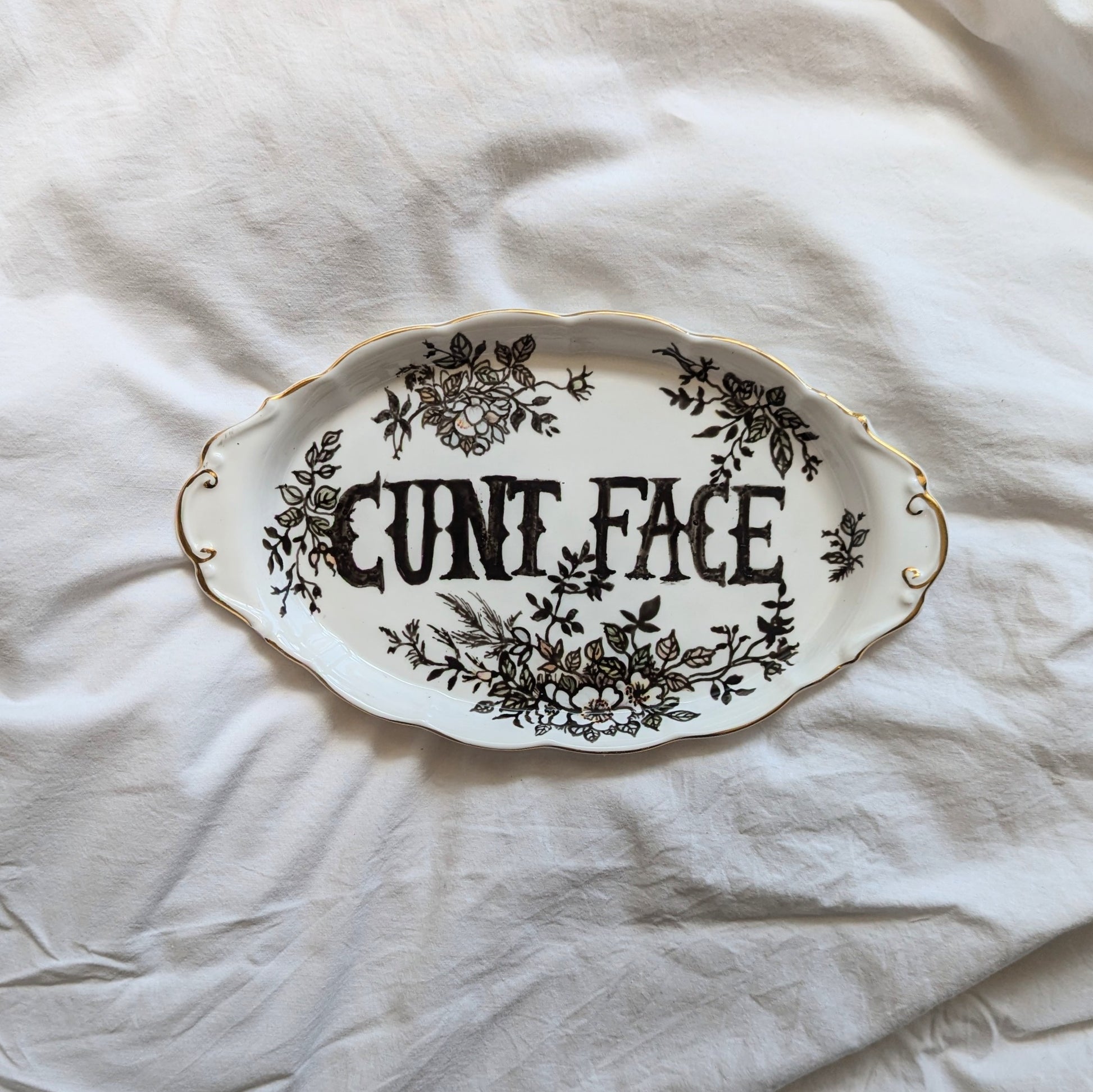 A decorative platter with some black outlined flora designs. In the centre in block gothic style font are the words "cunt face"