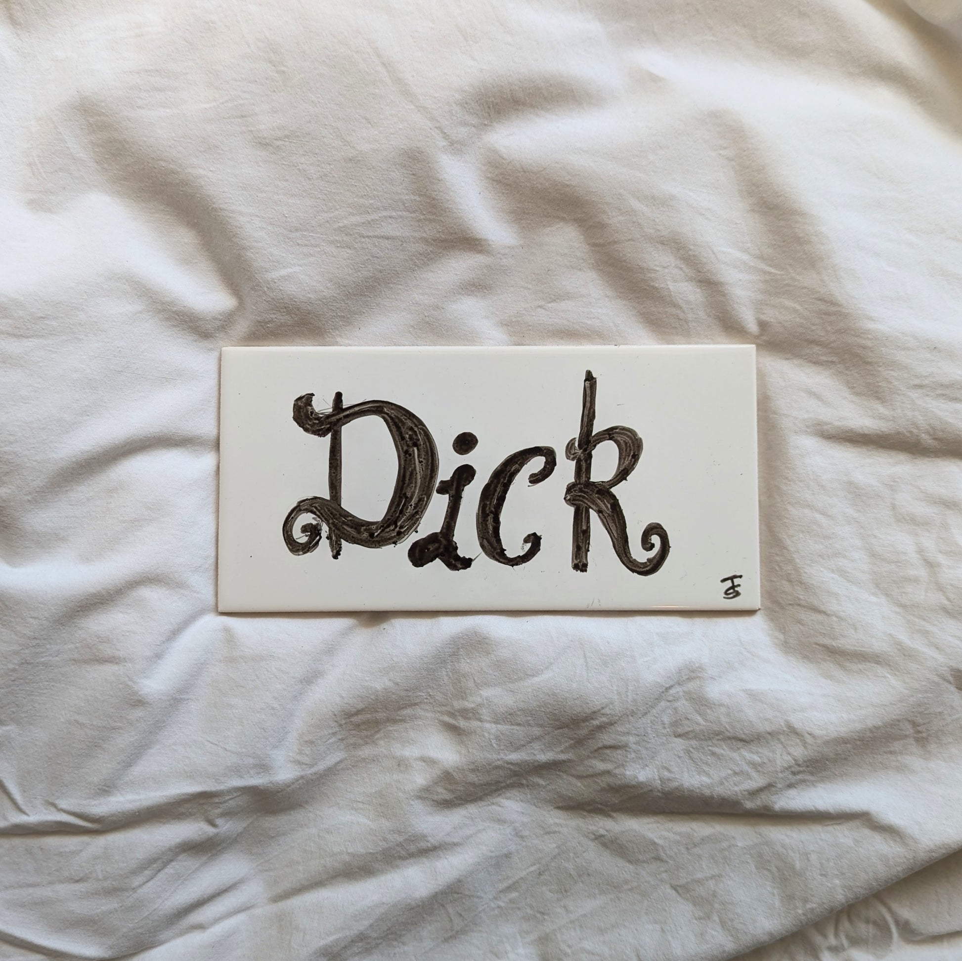 A ceramic tile with the word "dick" hand written in the centre in a cursive font. 