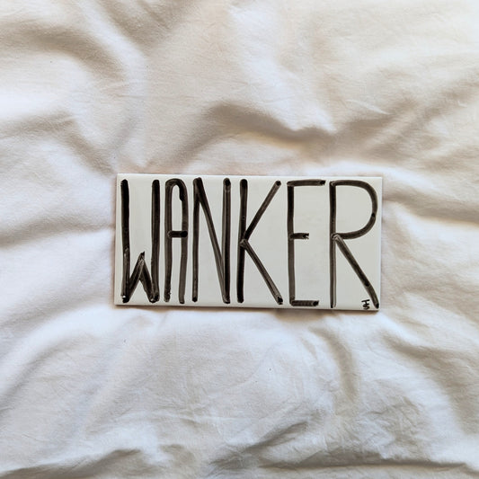 A small ceramic tile with the word "wanker" written in black ink in thin block letters filing the tile.