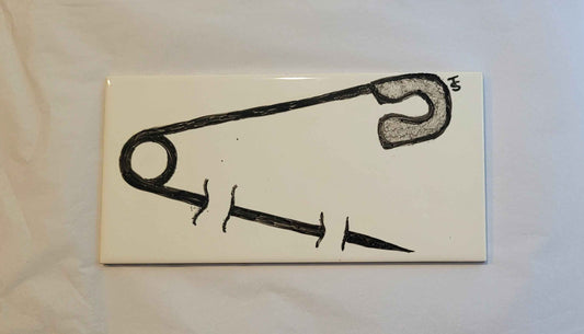 A ceramic tile featuring a hand painted ink drawing of a safety pin. The pin looks like it is pined through the tile. 
The pin is open. 