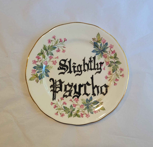 a small ceramic plate with a gold rim. the plate features a coloured print of small pink flowers and green foliage. it also has the words "slightly psycho" written by hand in a blackletter style font across two lines roughly in the middle. 