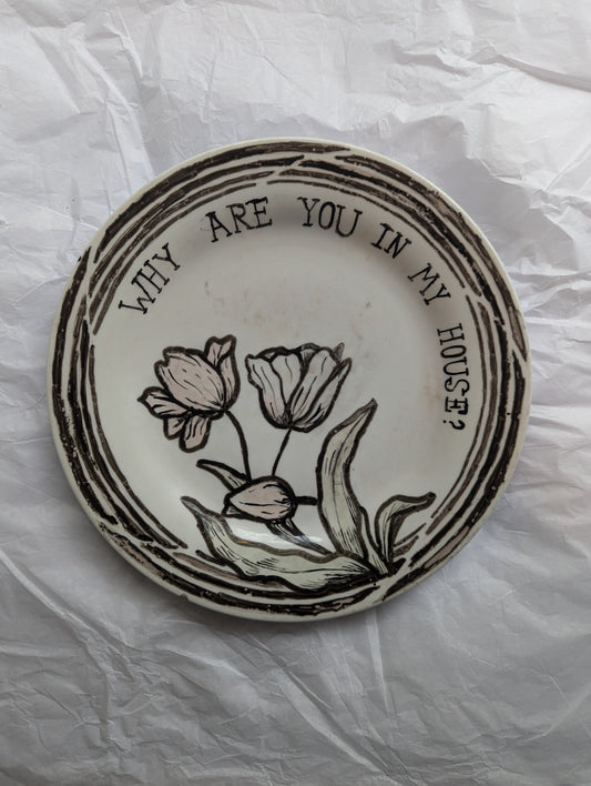 A large ceramic plate featuring a hand drawn ink outlined tulip and the word "why are you in my house?" written around the rim of the plate in a serif font. 